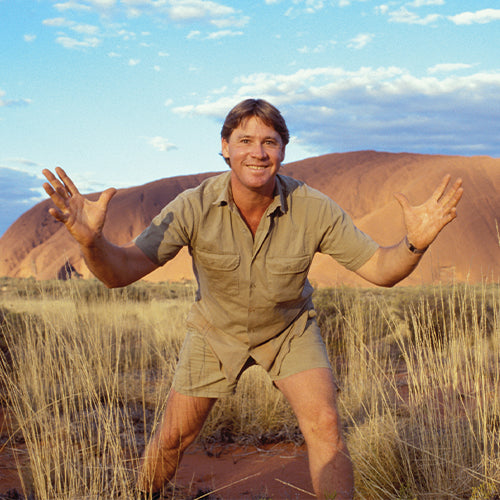 Steve Irwin in a safari outfit standing in a desert landscape with mountains in the background